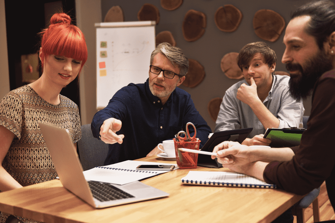 Team in an office having a meeting while one of the men is pointing at a laptop screen